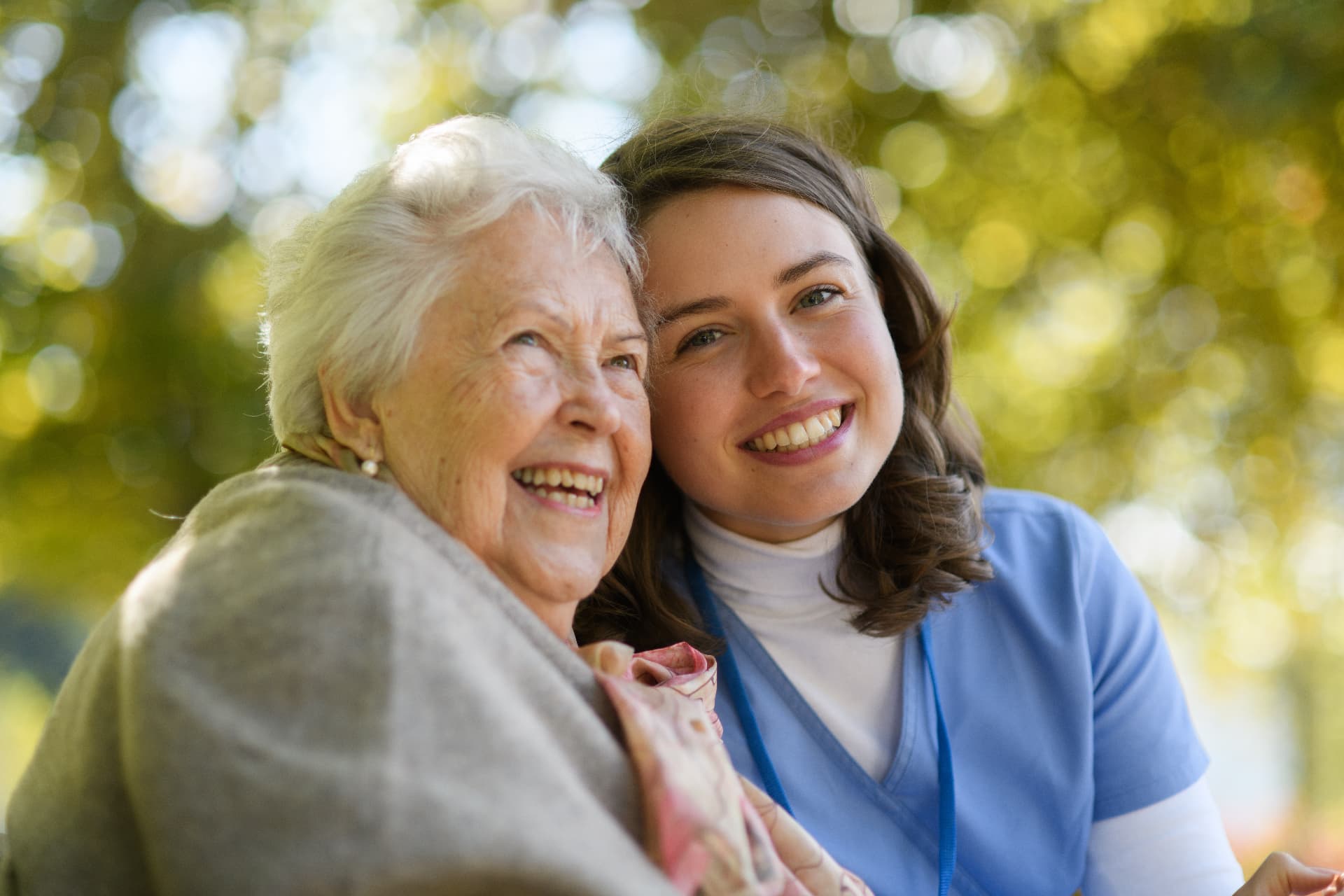 A support worker and an older woman smiling together outdoors beneath sunlit trees