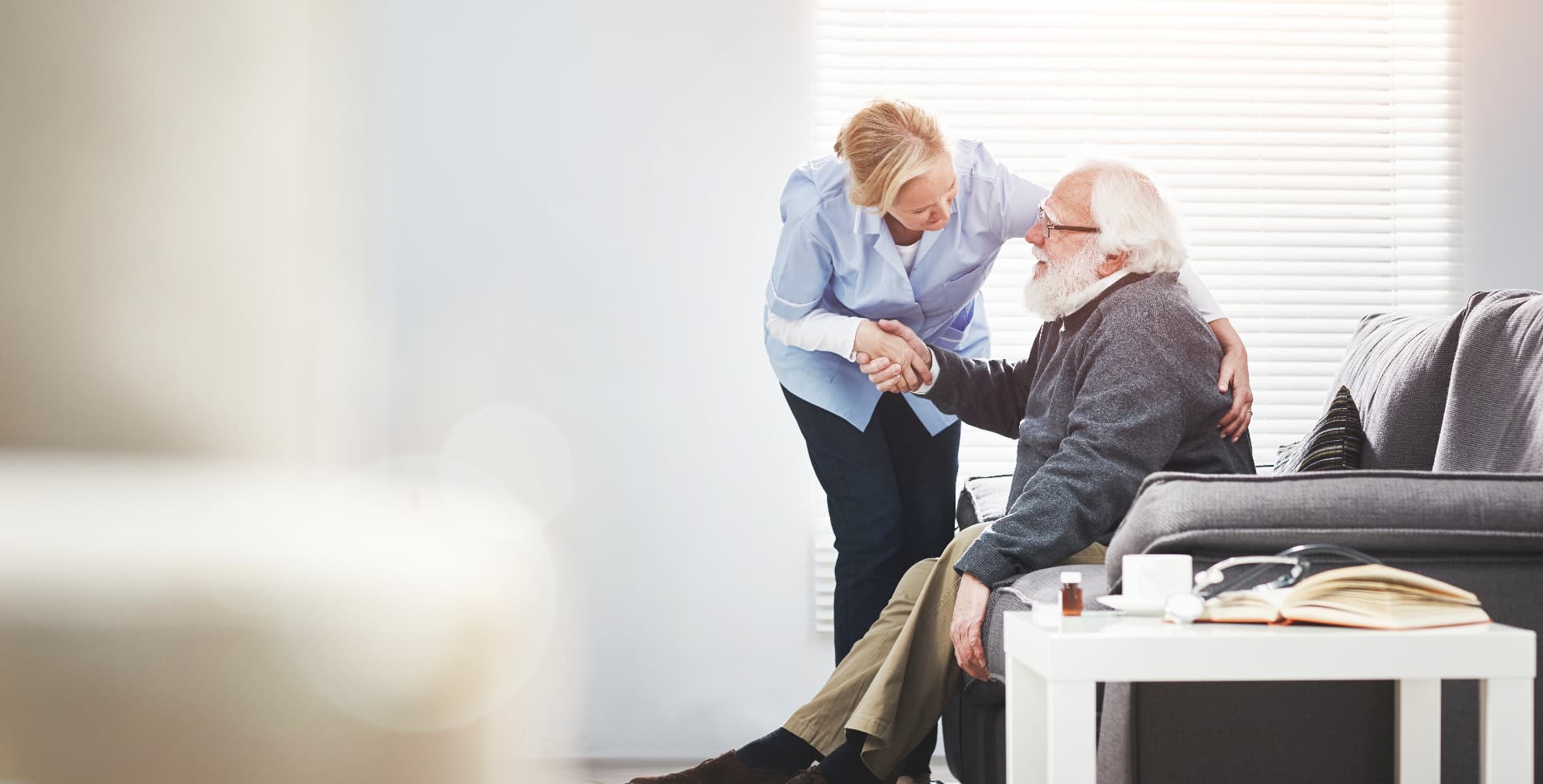 A clinician crouching beside an older man on a lounge, shaking his hand during a home visit