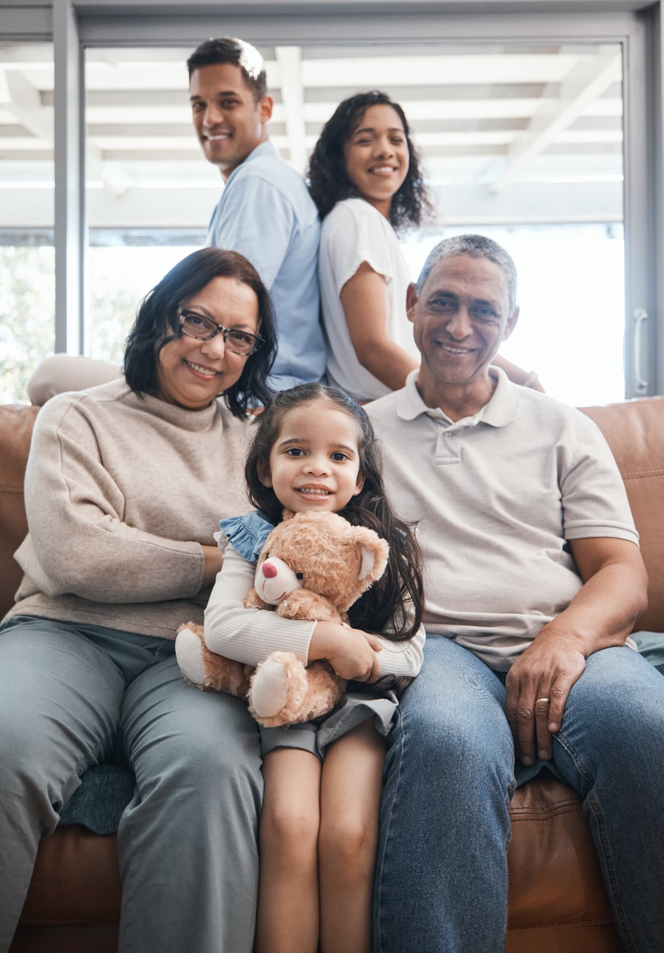 Three generations of a family sitting together on a lounge at home, a young girl holding a teddy bear