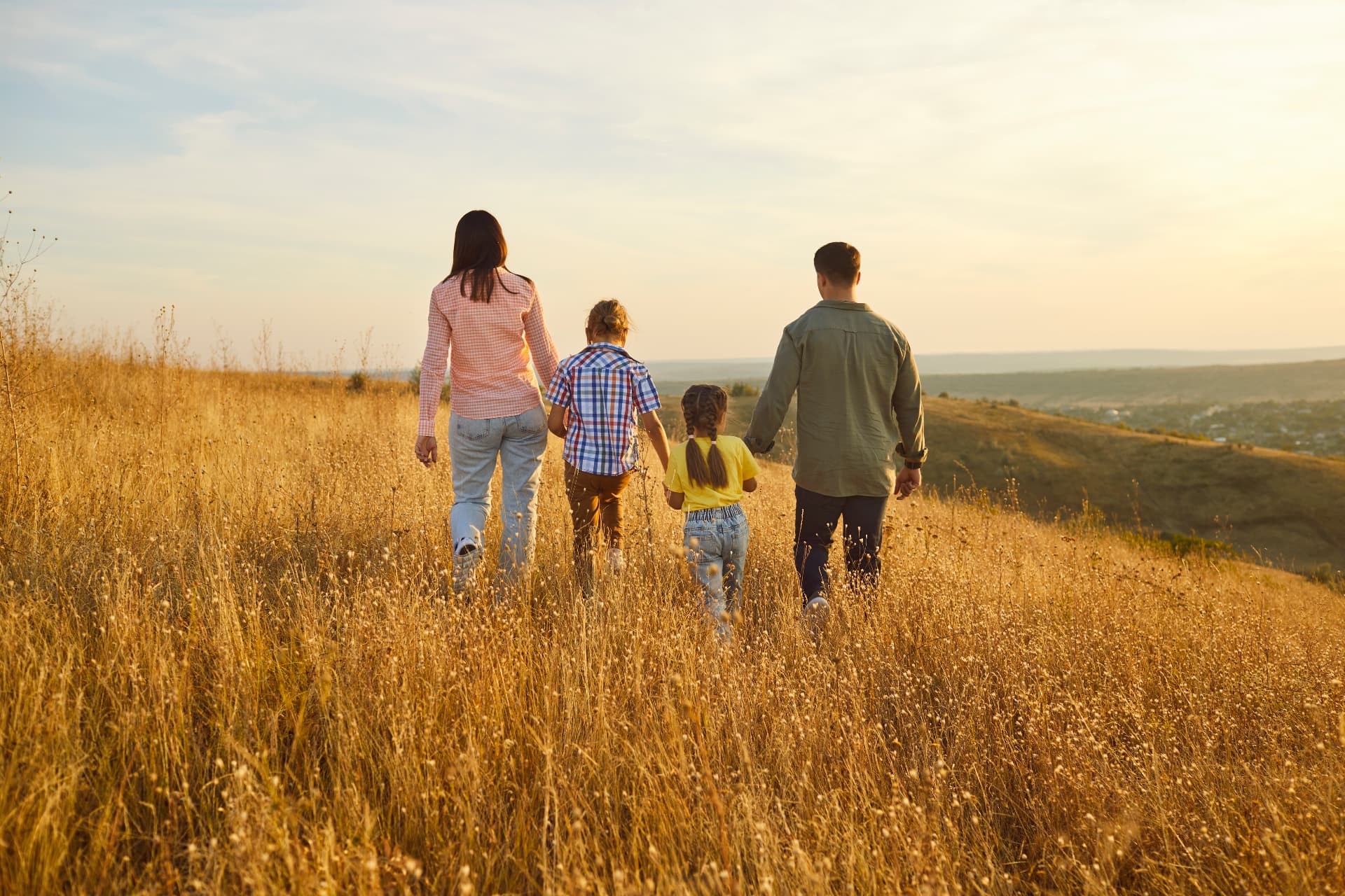 A family of four walking hand in hand through a sunlit grassy hillside at golden hour