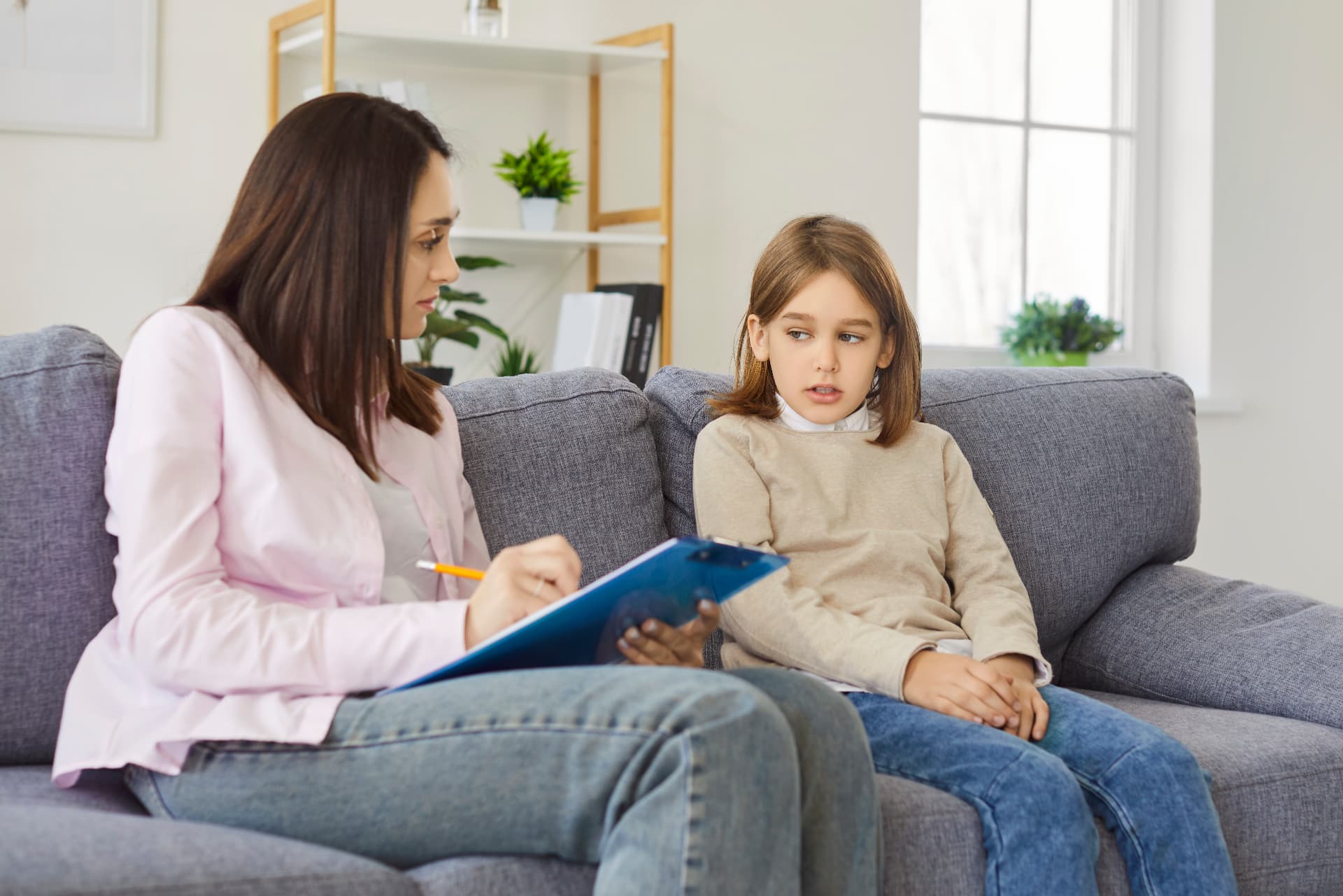 A clinician sitting on a lounge with a young girl, taking notes on a clipboard during a session